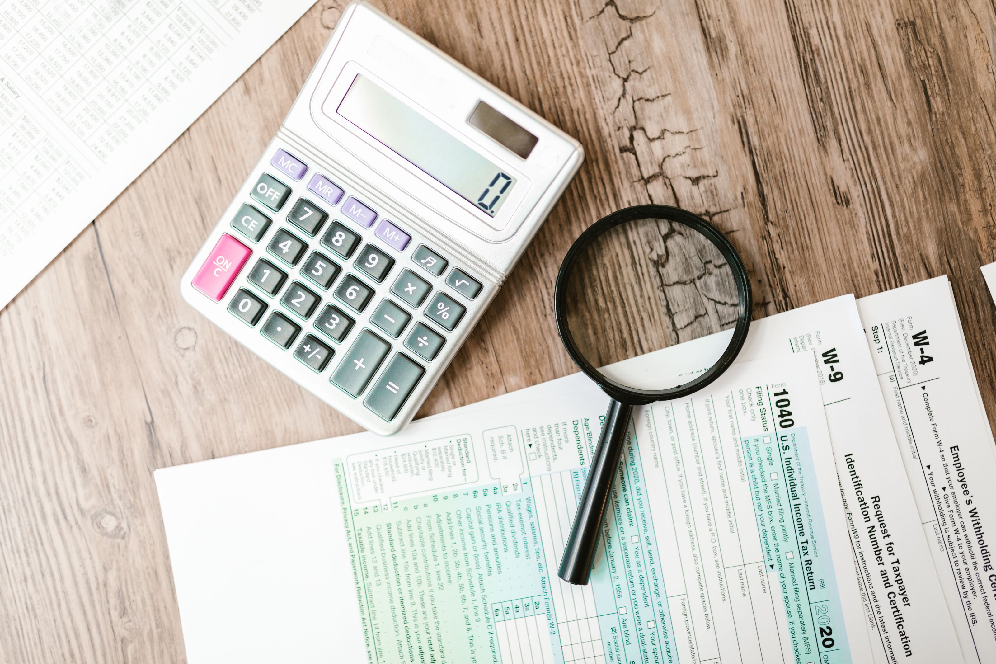 A calculator and magnifying glass beside US tax forms on a wooden desk.