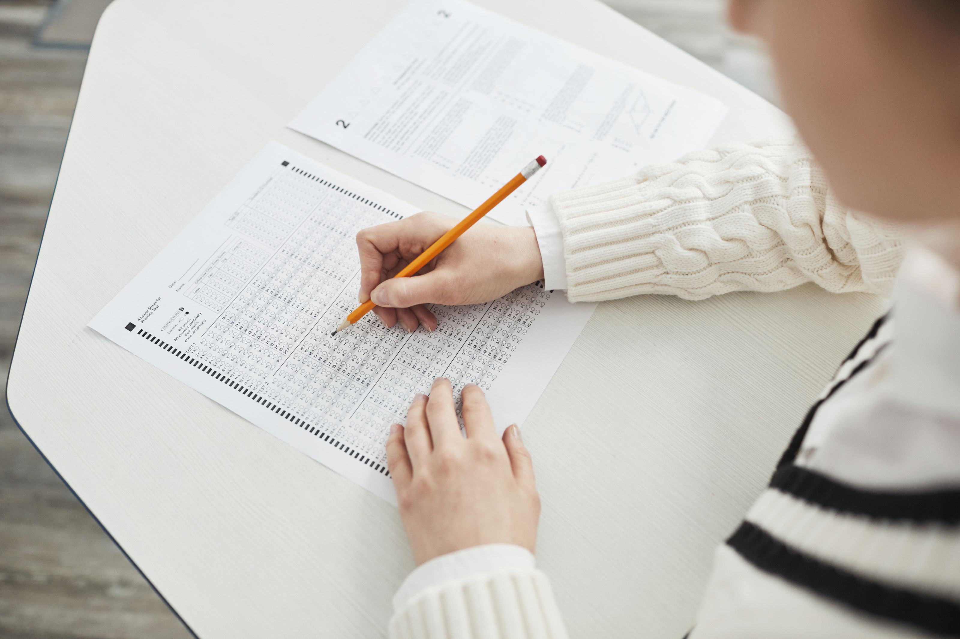 Close-up of a person in a cream sweater using a yellow pencil to fill in a multiple-choice answer sheet, with an exam paper on the desk.