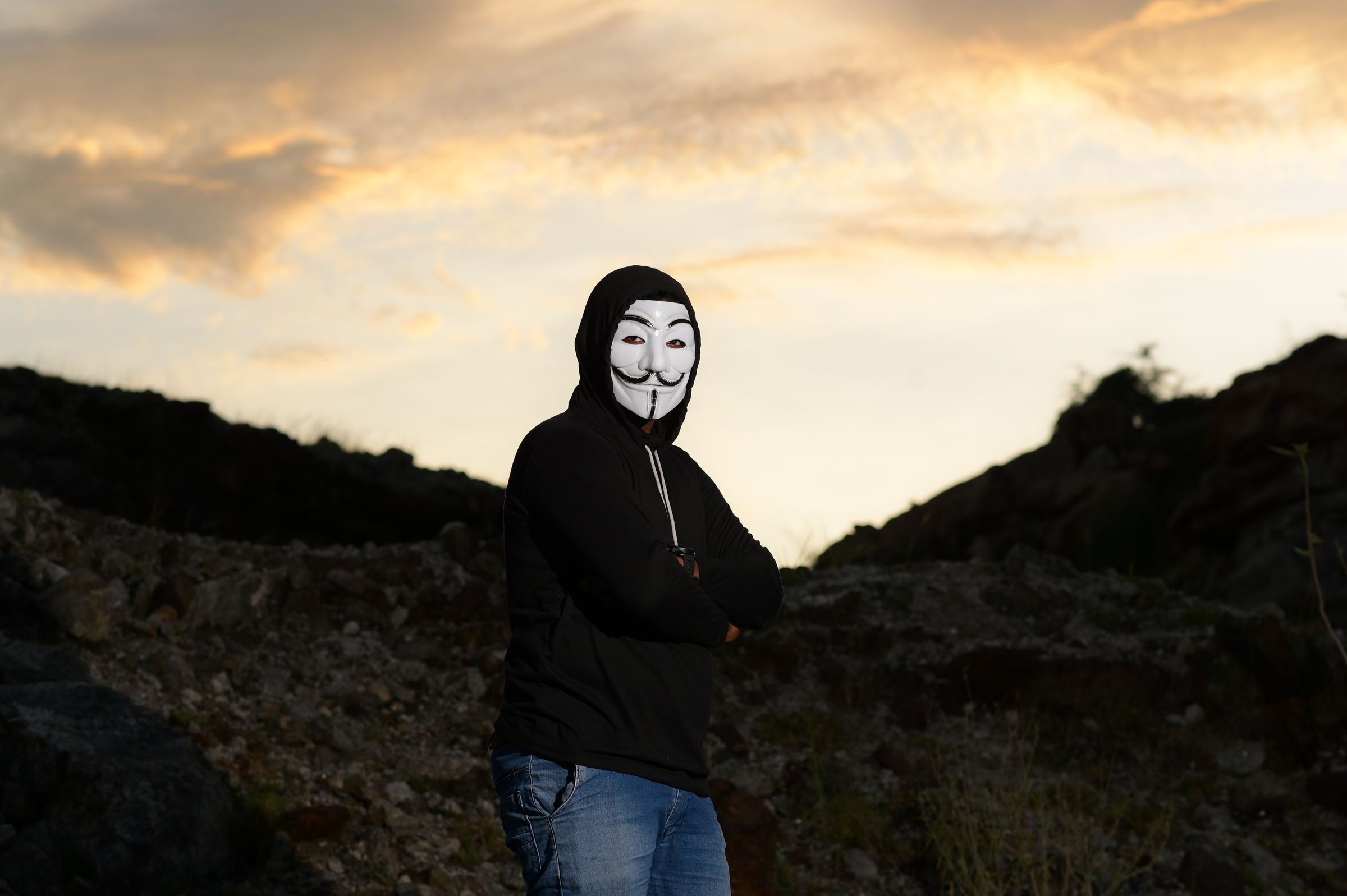 Silhouette of a person in a Guy Fawkes mask and hoodie standing outdoors during sunset.